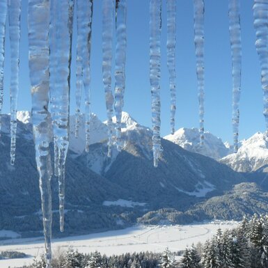 Eiszapfen im Vordergrund mit Blick auf eine winterliche Berglandschaft und ein verschneites Tal rund um den Bauernhof.