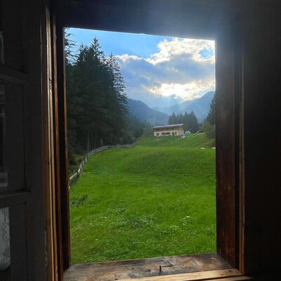 Ein idyllischer Blick aus dem rustikalen Holzfenster der Alm auf saftig grüne Wiesen und die umliegende Berglandschaft.