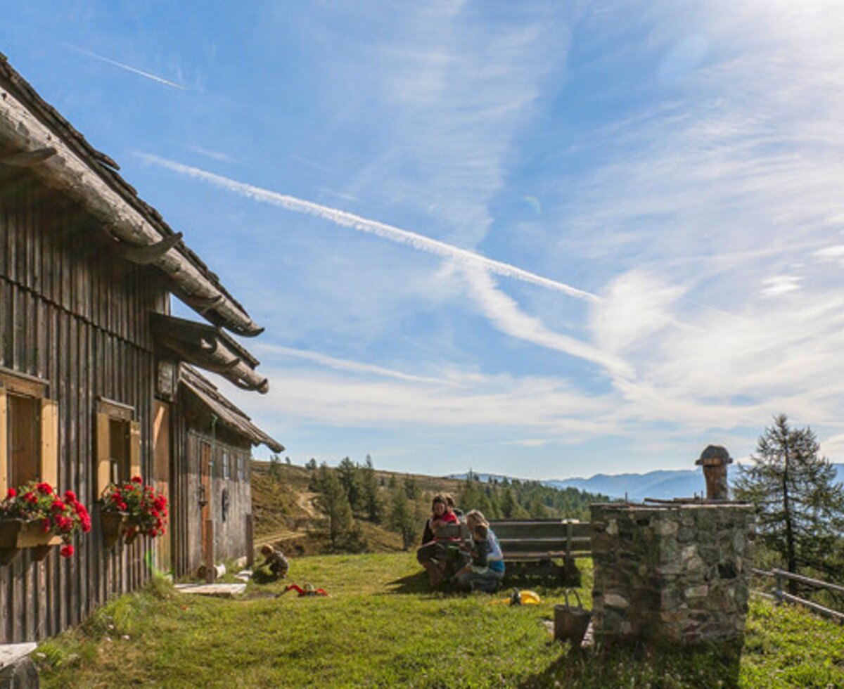 Die Alm mit Holzfassade, Blumenkästen und einem Außenbereich mit Sitzgelegenheiten sowie Bergblick.