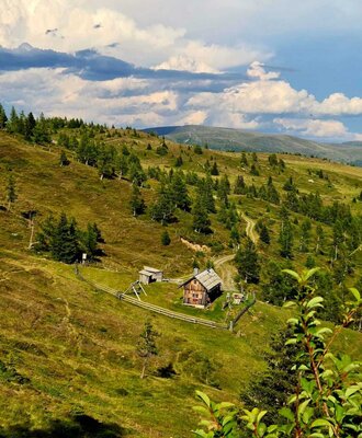Die urige Alm eingebettet in eine sanfte, grüne Berglandschaft unter einem weiten Wolkenhimmel.