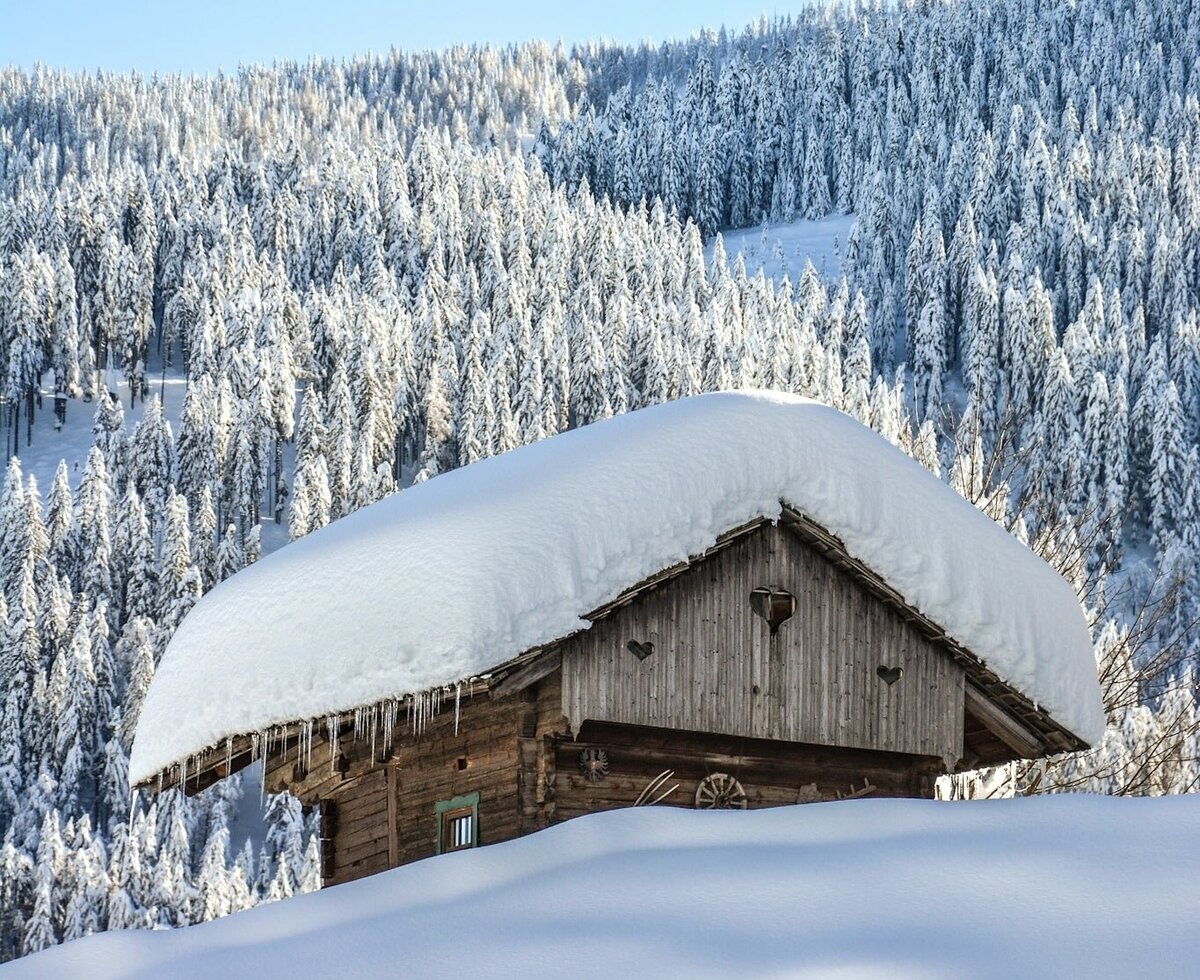Das Bauernhaus im Winter, mit einer dicken Schneedecke auf dem Dach, Eiszapfen und einem schneebedeckten Wald im Hintergrund.