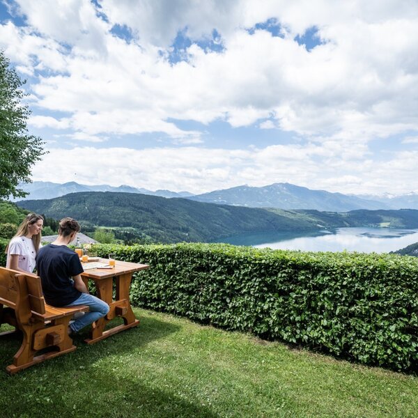 Sitzbereich im Freien am Ferienhaus mit Holztisch und Bank, mit Panoramablick auf den See und die Berge.