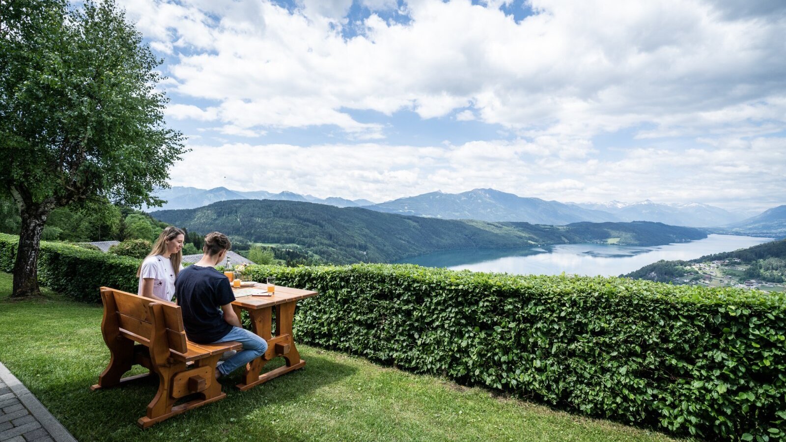 Sitzbereich im Freien am Ferienhaus mit Holztisch und Bank, mit Panoramablick auf den See und die Berge.