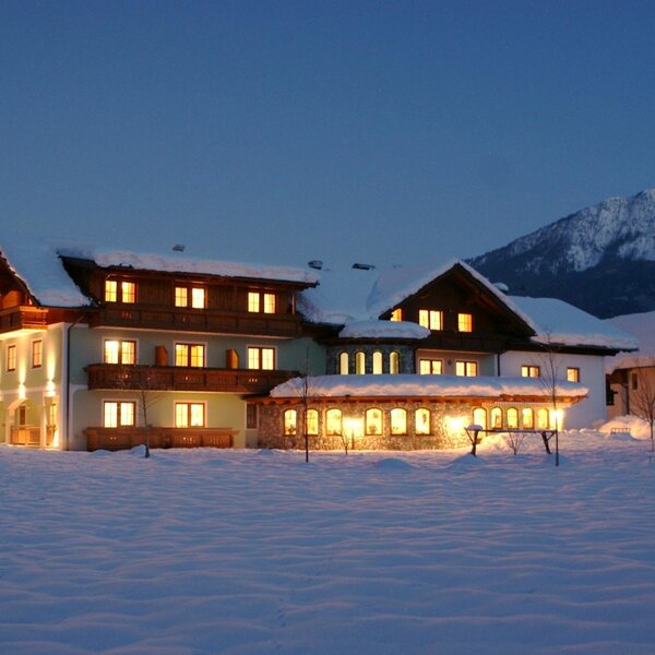 Der beleuchtete Bauernhof im Schnee bei Nacht, mit warm leuchtenden Fenstern und Balkonen vor der Kulisse der Berge.