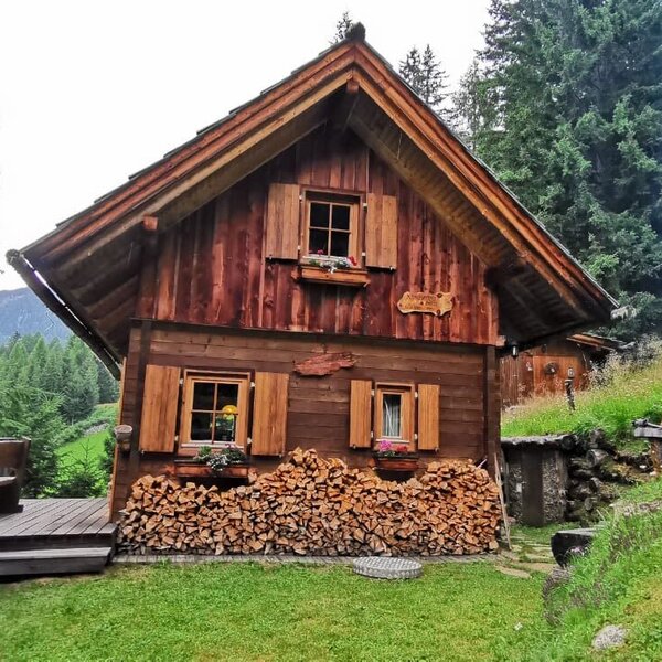 Außenansicht der Alm mit Holzfassade, Fensterläden, Blumenkästen, Holzstapel und einer kleinen Holzterrasse.