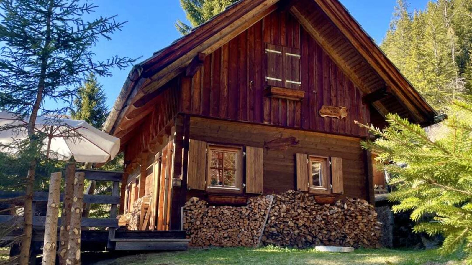 Die Alm von außen, mit Holzfassade, Giebeldach, Fensterläden und einem Holzstapel vor einer Rasenfläche.