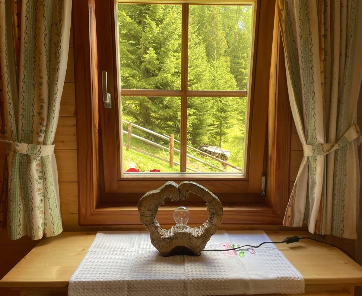Ein gemütlicher Ausblick aus dem Holzfenster der Alm auf den saftig grünen Wald, geschmückt mit einer rustikalen Tischlampe.