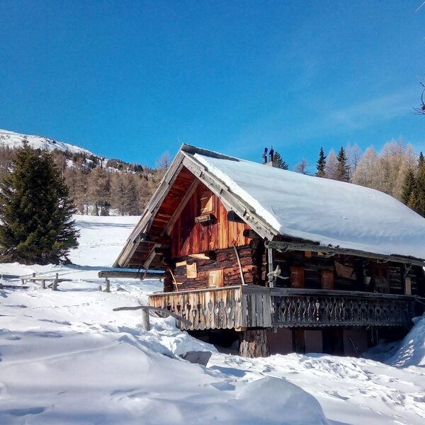 Die verschneite Alm mit Holzbalkon vor der winterlichen Bergkulisse.