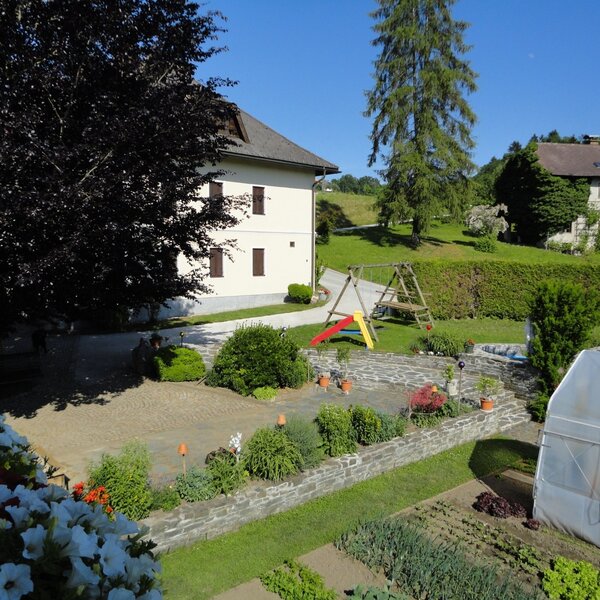 Der Außenbereich des Bauernhofs mit Kinderspielplatz, bepflanzten Beeten, Gewächshaus und einer gepflasterten Terrasse.
