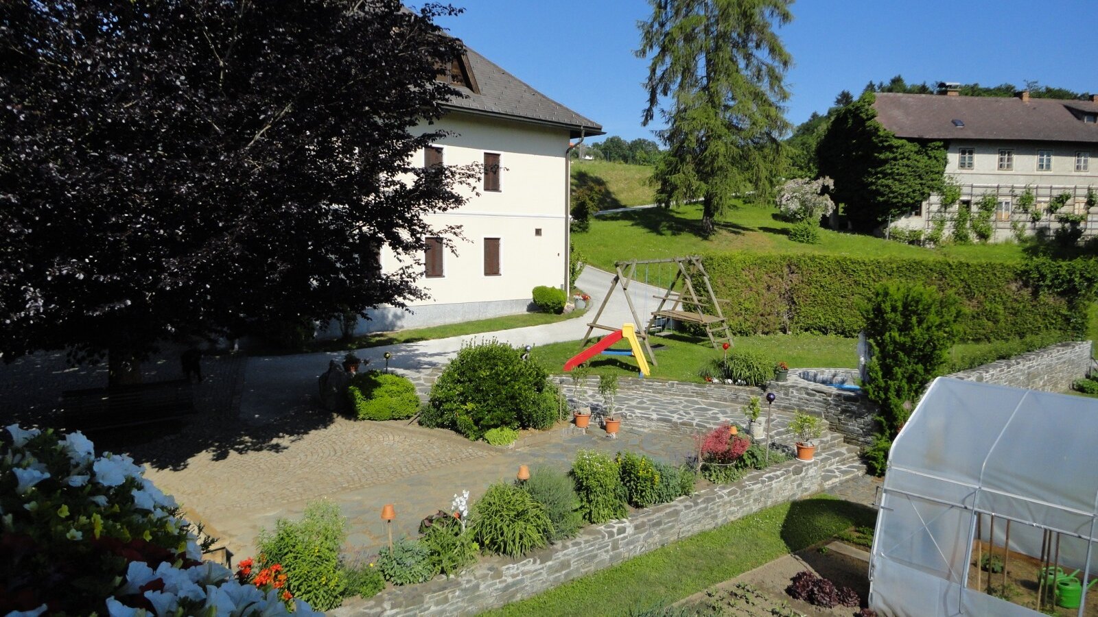 Der Außenbereich des Bauernhofs mit Kinderspielplatz, bepflanzten Beeten, Gewächshaus und einer gepflasterten Terrasse.