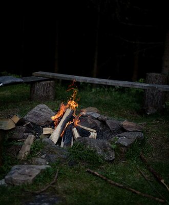 Lagerfeuerstelle auf der Alm mit brennendem Holz und einer Holzbank im Hintergrund.