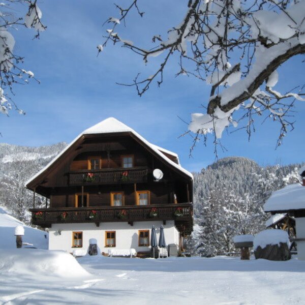 Der Bauernhof im Winter, mit schneebedecktem Gebäude und Balkonen, umgeben von verschneiten Bäumen und Bergen.
