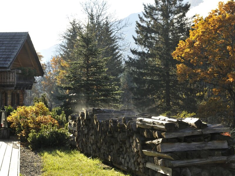 Der Bauernhof mit gestapeltem Brennholz neben dem Gebäude, umgeben von Wald und Bergen.