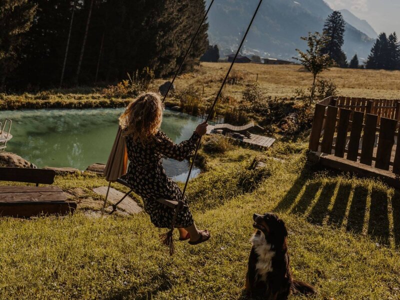 Der Außenbereich des Bauernhofs mit einem Naturschwimmteich, einer Schaukel und einem Hund, umgeben von grüner Landschaft und Bergen.