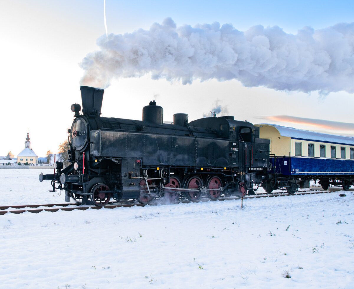 Eine schwarze Dampflokomotive mit weißem Rauch und Personenwagen auf verschneiten Gleisen, mit einer Kirche im Hintergrund, in der Umgebung des Bauernhofs.
