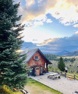 Die Alm mit einer großen Holzterrasse und Sitzgelegenheiten im Freien, umgeben von bewaldeten Bergen.