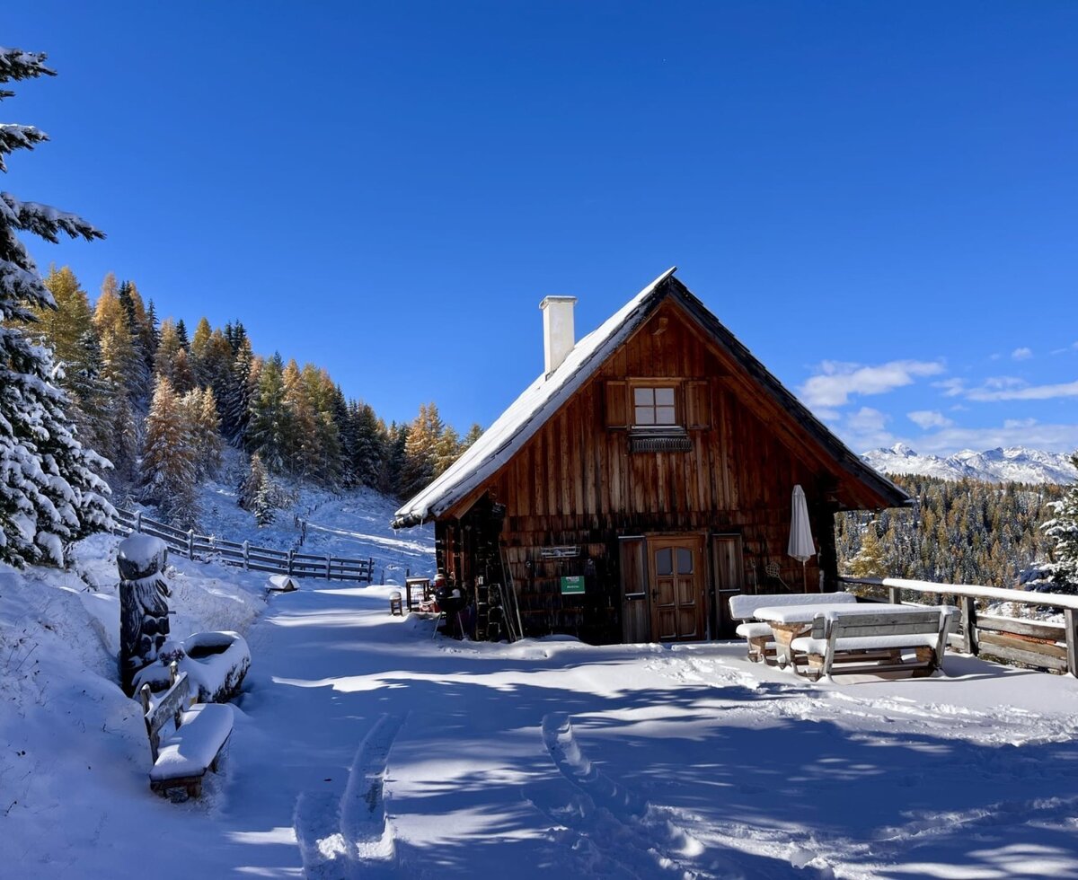 Die Alm im Winter, umgeben von Schnee und Bergen, mit einem verschneiten Holztisch und Bänken davor.