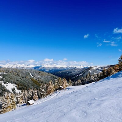 Winterlandschaft mit verschneiten Bergen, Wäldern und Almhütten unter blauem Himmel.