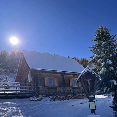 Die verschneite Alm unter blauem Himmel an einem sonnigen Wintertag, mit einem Wegkreuz im Vordergrund.