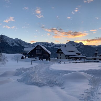 Der Bauernhof im Winter mit schneebedeckten Gebäuden und Feldern, umgeben von Bergen unter einem farbigen Himmel.