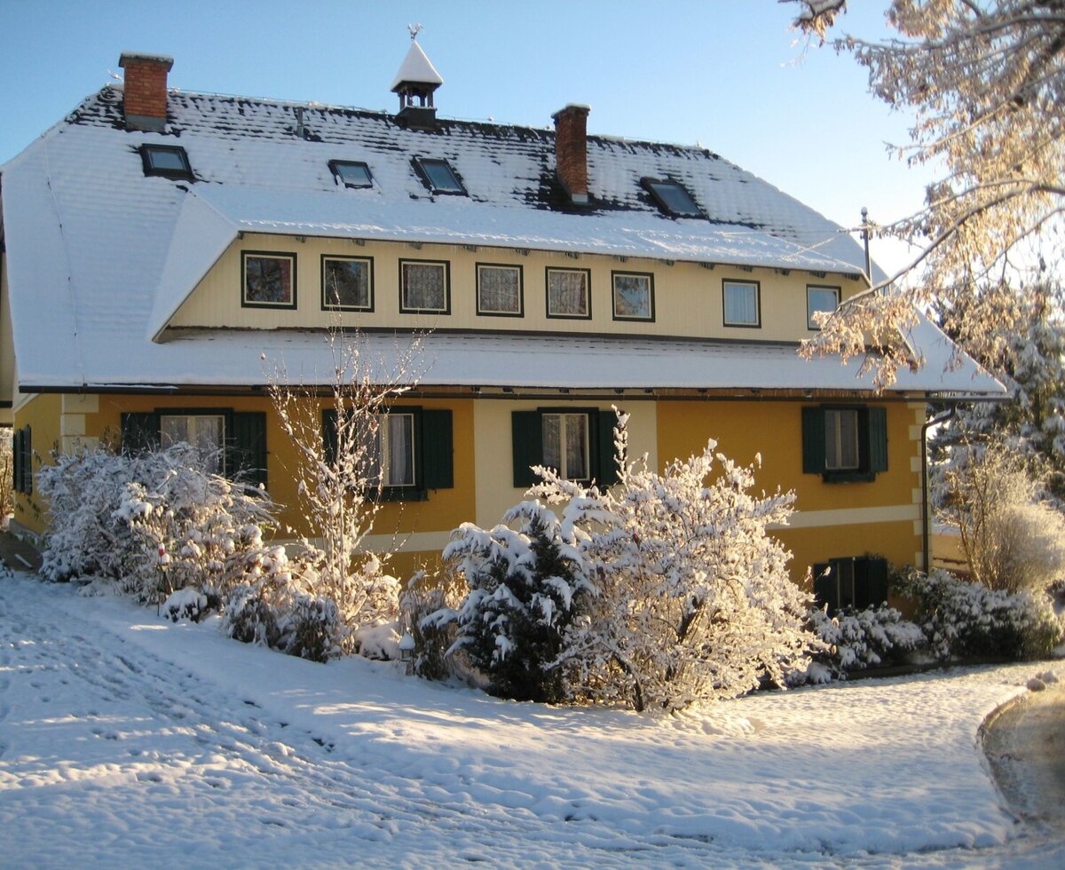 Die Außenansicht des Bauernhofs im Winter, mit gelber Fassade, grünen Fensterläden und schneebedecktem Dach.