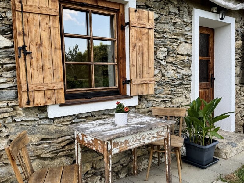 Außenbereich des Bauernhofs mit Steinfassade, Holzfensterläden und Sitzgelegenheiten.