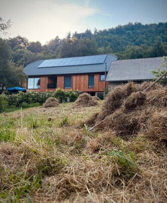 Der Bauernhof mit Holzfassade und Solarpaneelen auf dem Dach, gelegen an einem Hang mit Wald im Hintergrund.