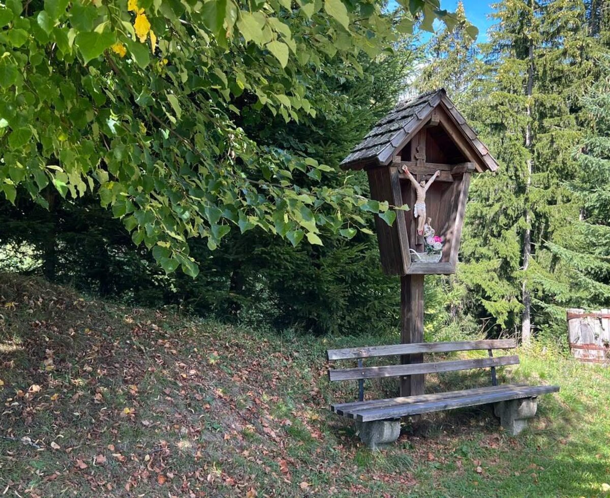 Ein friedlicher Rastplatz am Bauernhof mit einer rustikalen Holzbank und einem traditionellen Hofkreuz am Waldrand.