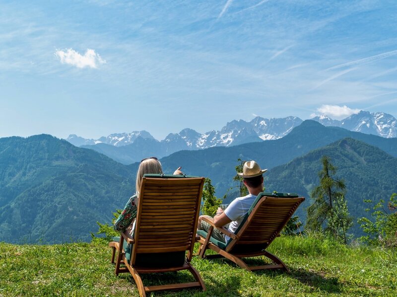 Die Alm bietet einen Panoramablick auf die Berge mit schneebedeckten Gipfeln, genossen von zwei Holzliegen auf der Wiese.