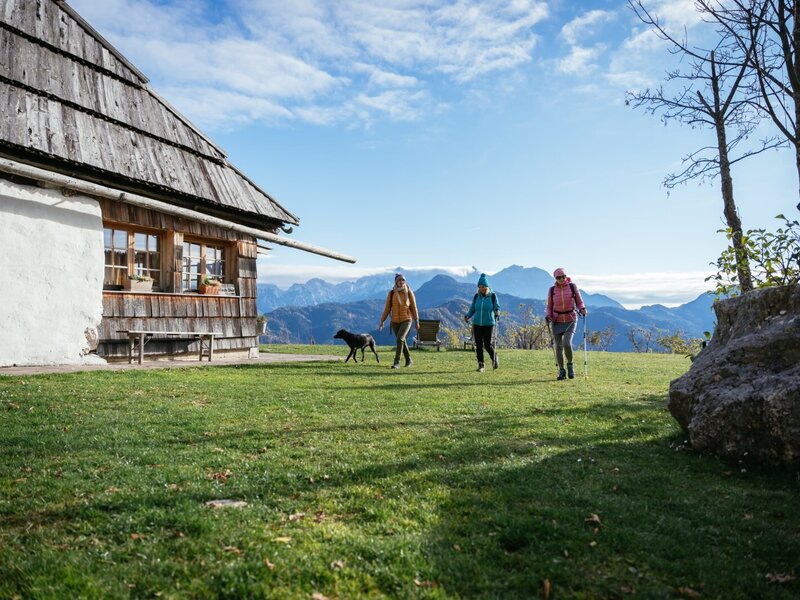 Das Gebäude des Hofs mit einer Gruppe Wanderer und einem Hund auf der Wiese und Blick auf die Berge.