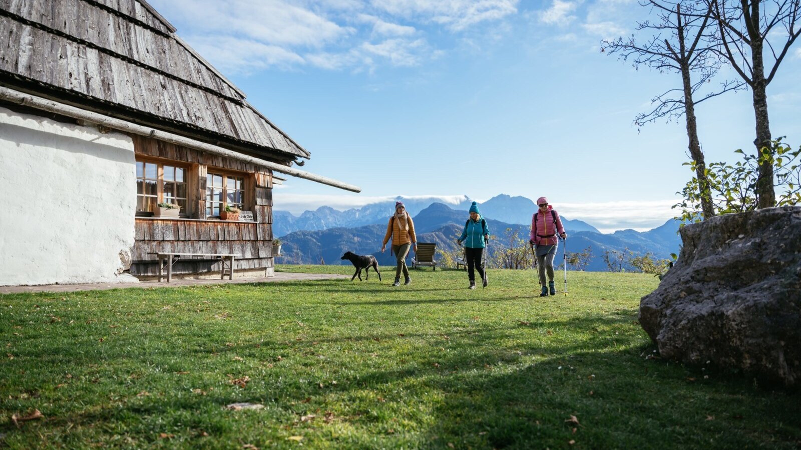 Das Gebäude des Hofs mit einer Gruppe Wanderer und einem Hund auf der Wiese und Blick auf die Berge.