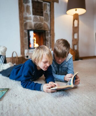 Zwei Kinder lesen auf einem Teppich in der Alm vor einem Steinkamin.
