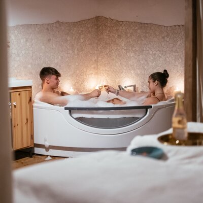 Der Whirlpool für zwei Personen in der Alm, ergänzt durch eine Natursteinwand und eine Liege im Vordergrund.