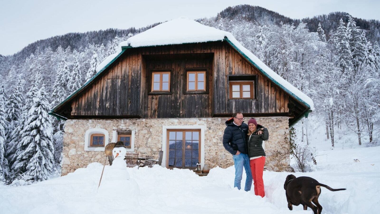 Die Alm in verschneiter Winterlandschaft mit einer Fassade aus Holz und Stein, einem Schneemann und einem schneebedeckten Bergwald im Hintergrund.