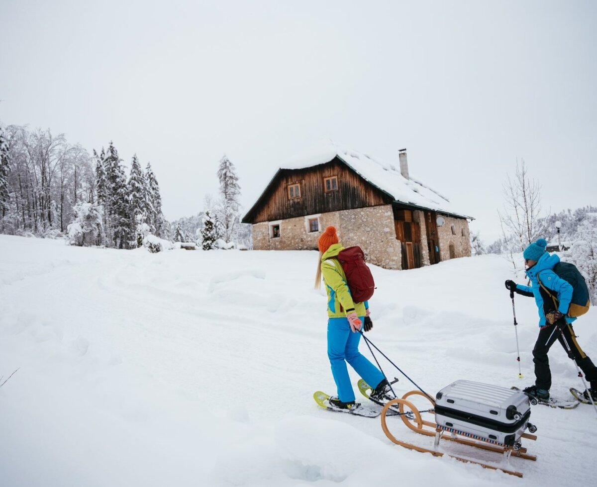 Zwei Personen wandern im Winter mit Schneeschuhen zur Alm und ziehen ihr Gepäck auf einem Schlitten.