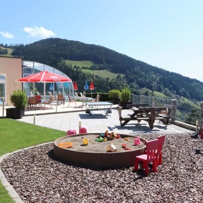 Außenbereich der Ap­par­te­ments mit Rasenfläche, Kinderspielplatz, Terrasse mit Sitzgelegenheiten, Tischtennisplatte, Poolbereich und Bergblick.