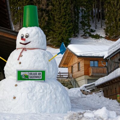 Ein großer Schneemann mit dem "Urlaub am Bauernhof"-Schild vor dem winterlichen Bauernhof.