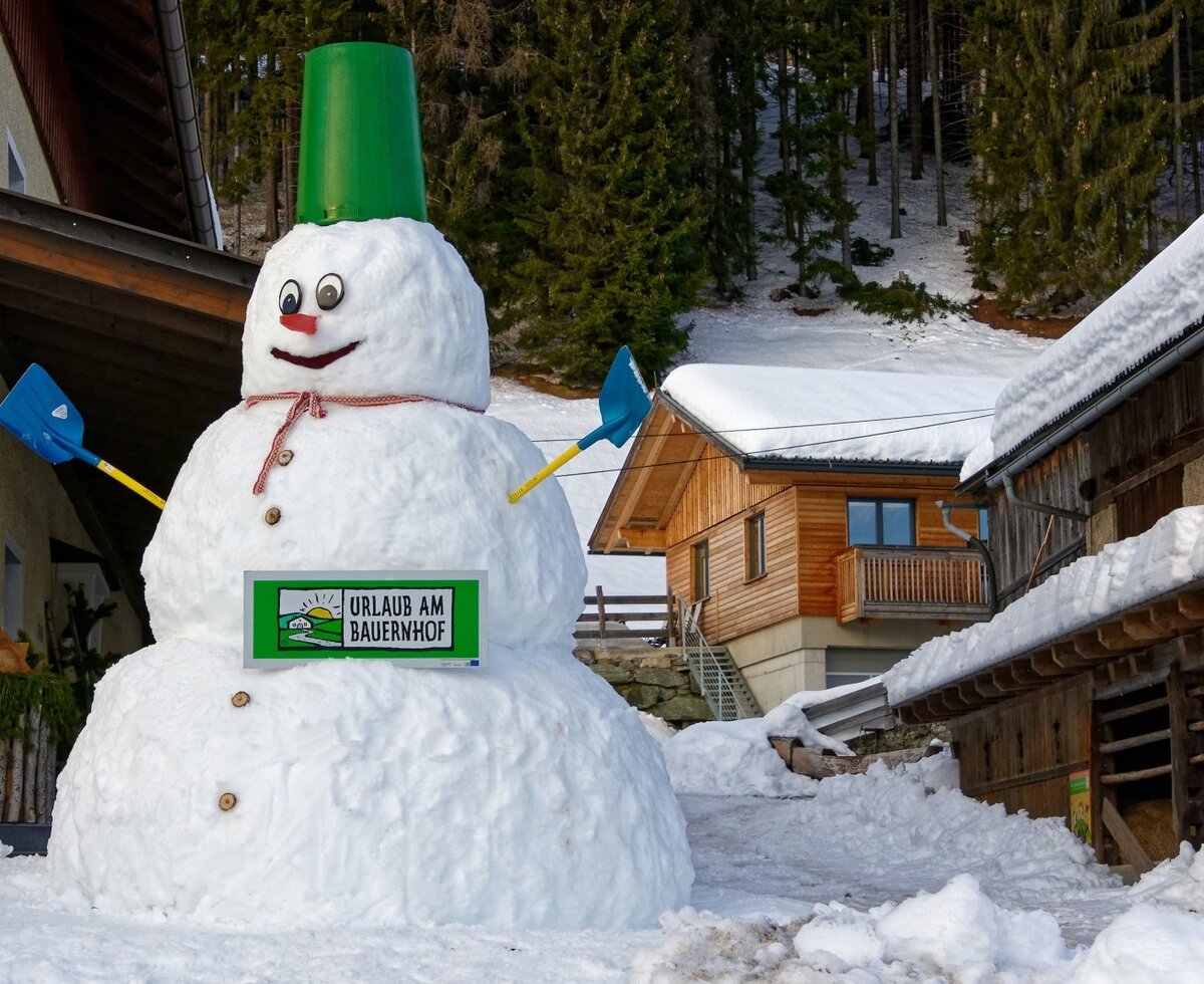 Ein großer Schneemann mit dem "Urlaub am Bauernhof"-Schild vor dem winterlichen Bauernhof.