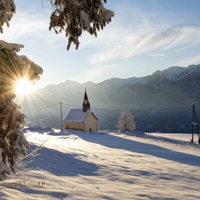 Die verschneite Winterlandschaft rund um den Bauernhof, mit einer kleinen Kirche und Bergen im Sonnenlicht.