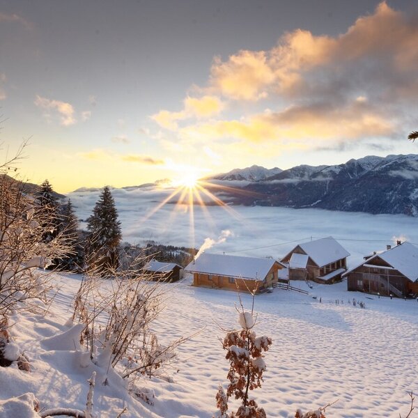 Der Bauernhof in einer verschneiten Winterlandschaft mit Sonnenaufgang über dem Tal und den Bergen.