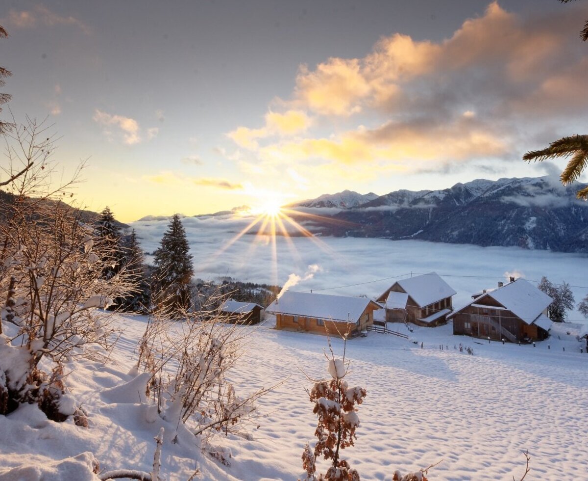Der Bauernhof in einer verschneiten Winterlandschaft mit Sonnenaufgang über dem Tal und den Bergen.