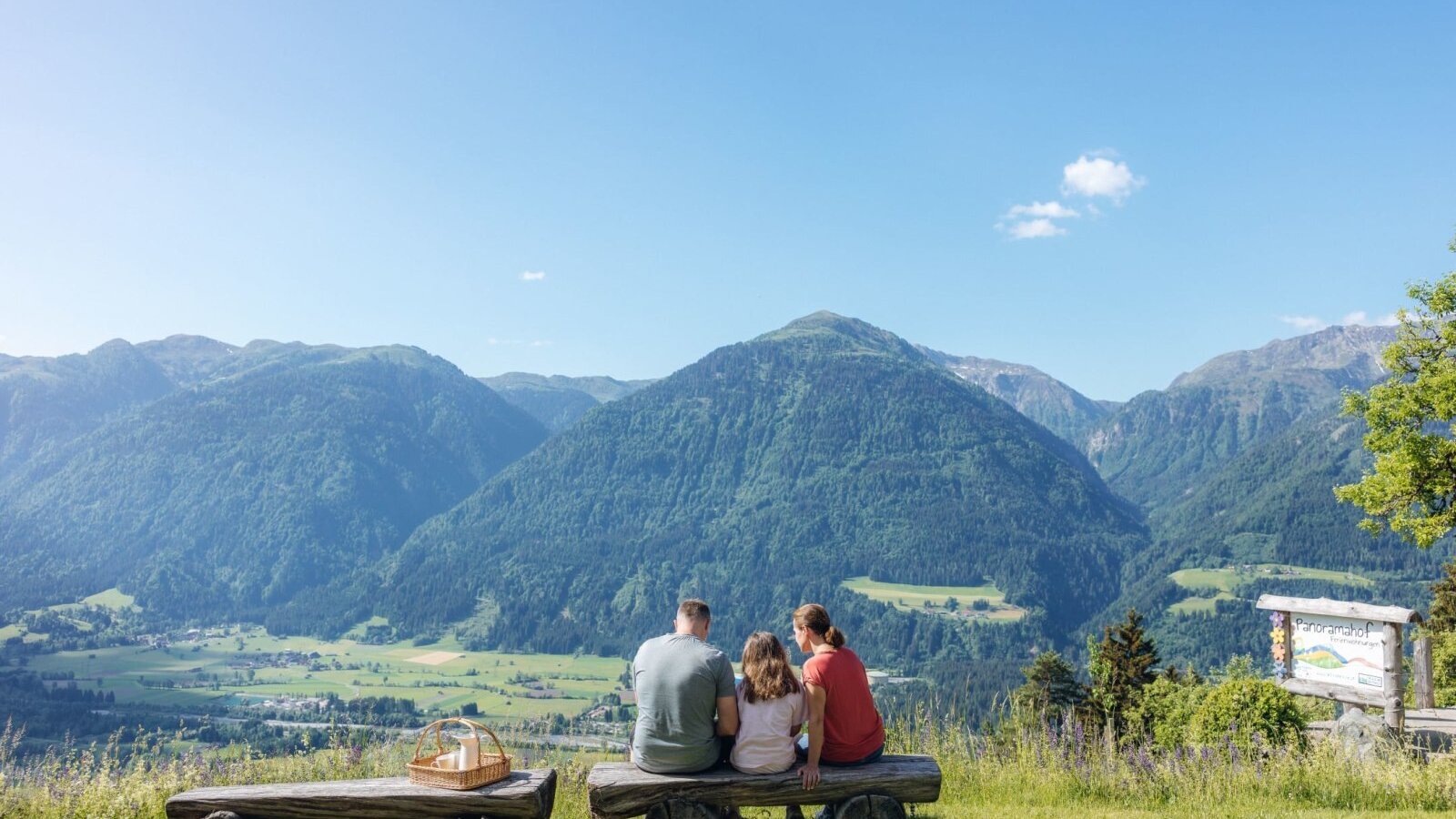 Weitläufiger Bergblick vom Bauernhof, genossen von einer Familie auf einer Holzbank mit Picknickkorb.