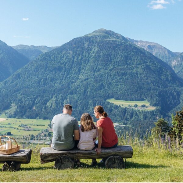 Eine Familie genießt auf Holzbänken mit einem Picknickkorb den Panoramablick auf die Berge und das Tal vom Bauernhof Panoramahof.