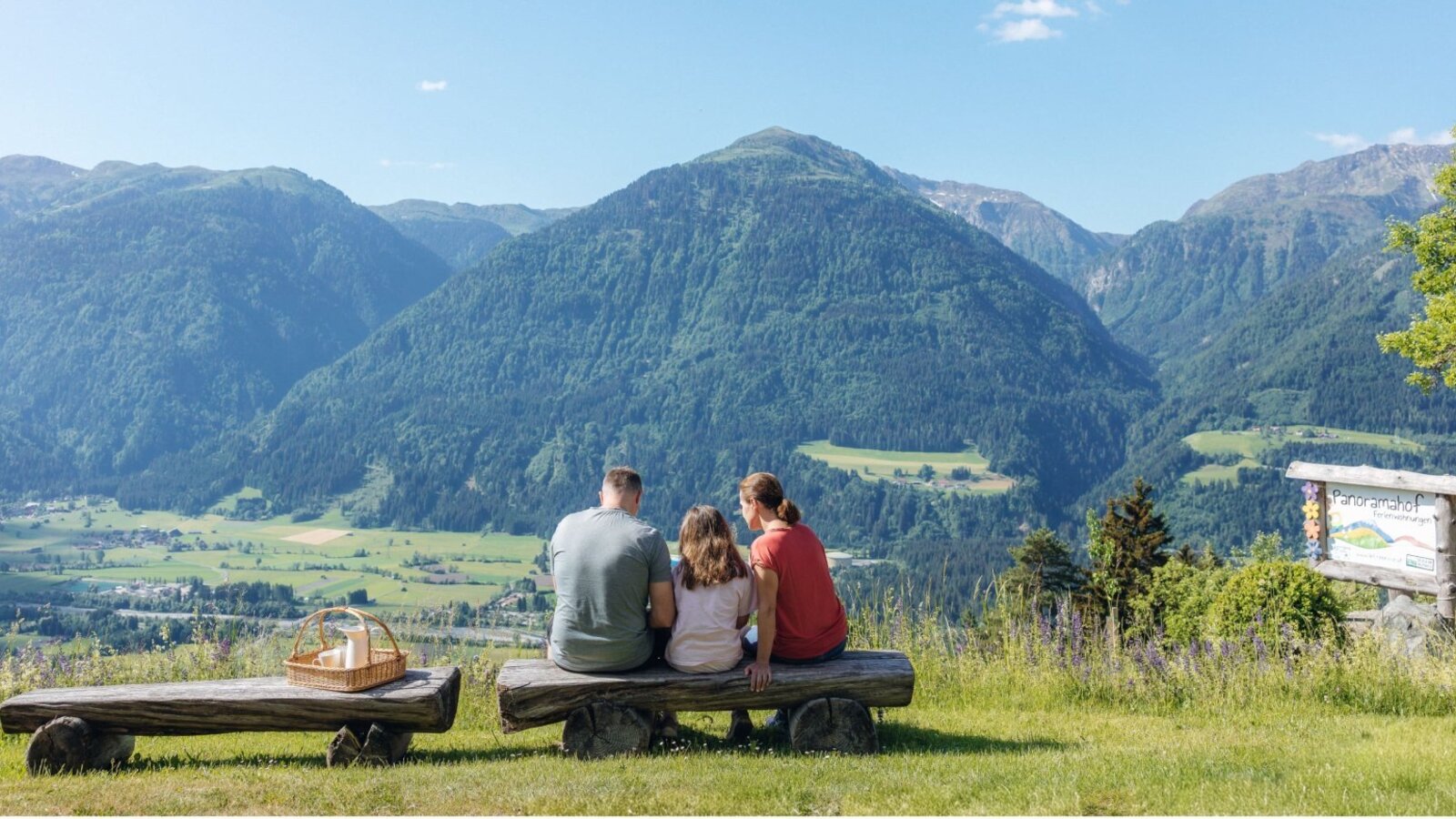 Eine Familie genießt auf Holzbänken mit einem Picknickkorb den Panoramablick auf die Berge und das Tal vom Bauernhof Panoramahof.