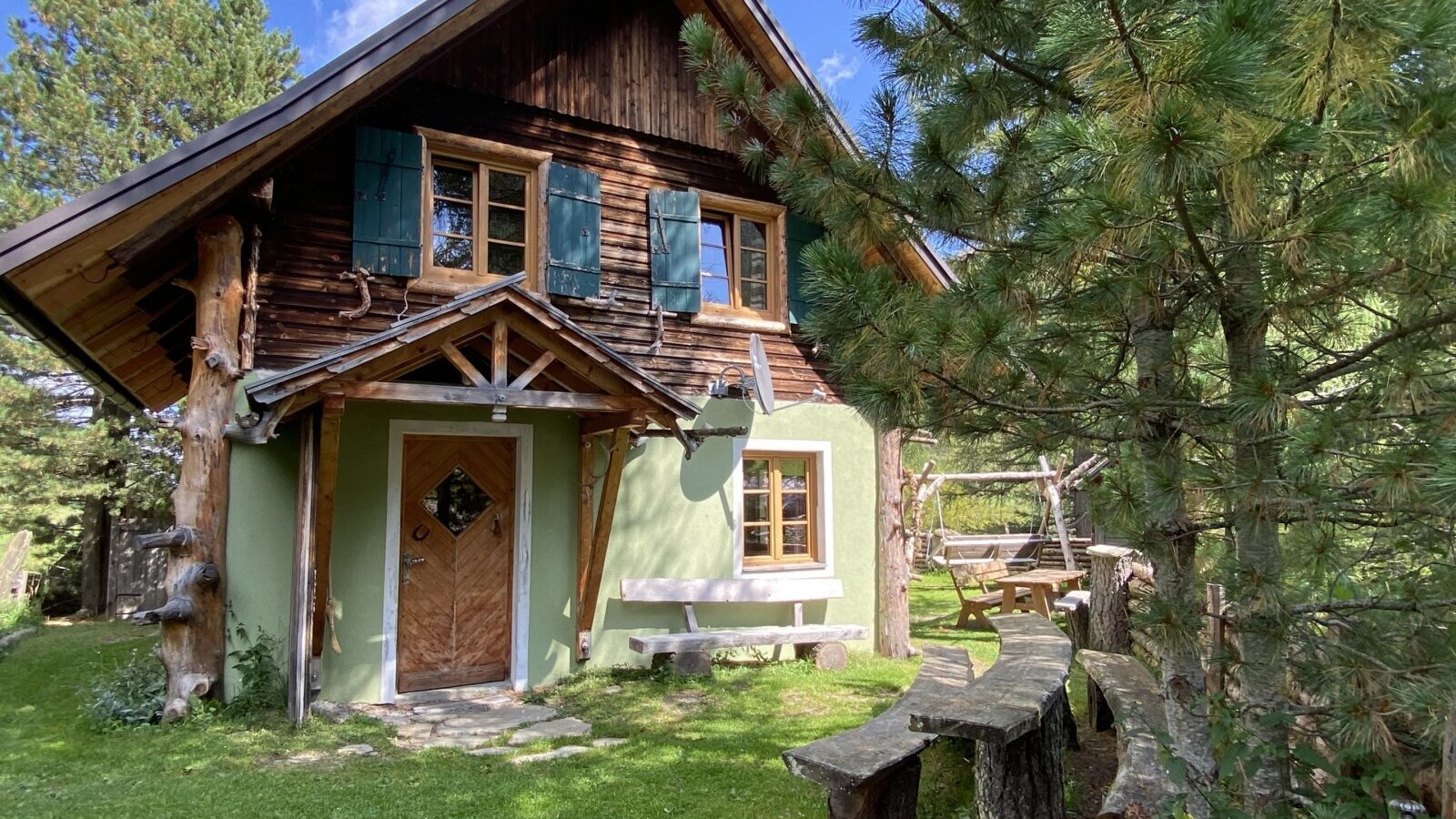 Die Alm von außen mit Holzfassade, grünen Wänden und einem Außenbereich mit Holzbänken und einem Tisch.