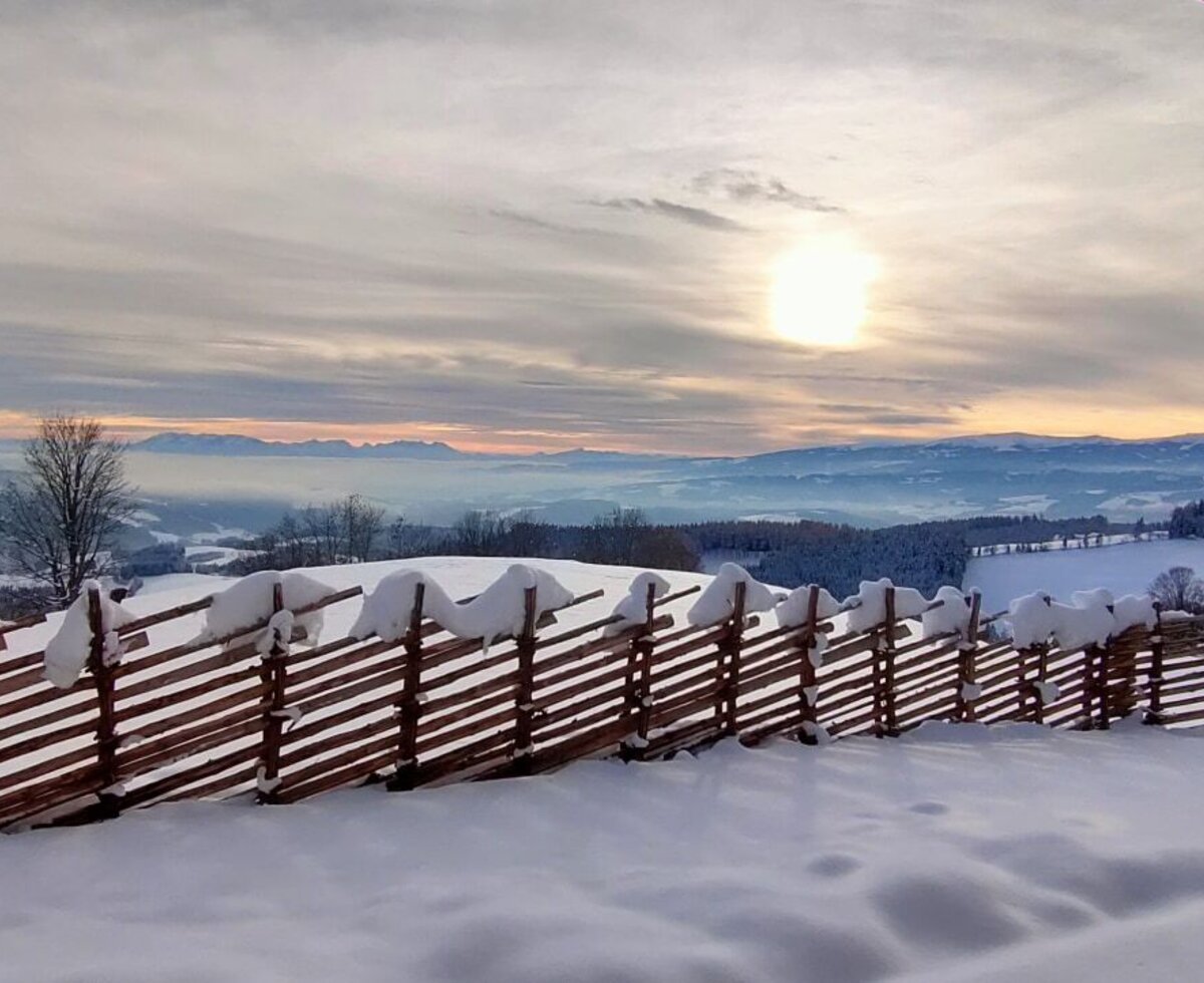 Blick von der Alm auf die verschneite Winterlandschaft mit Bergen und einem traditionellen Holzzaun.