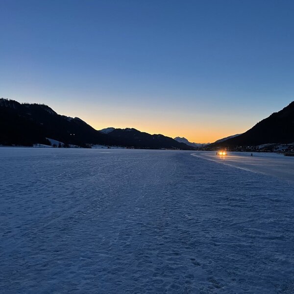 Die winterliche Landschaft mit gefrorenem See, umliegenden Bergen und entfernten Lichtern bei Dämmerung.