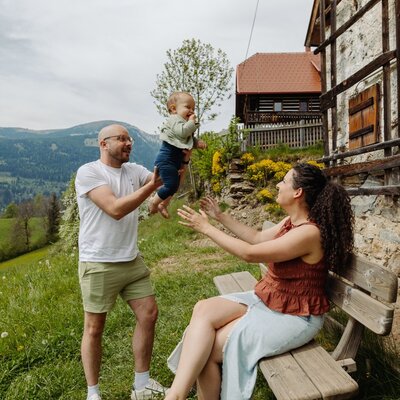 Außenbereich des Bauernhofs mit Rasenfläche, Holzbank und traditionellen Gebäuden vor Bergkulisse.