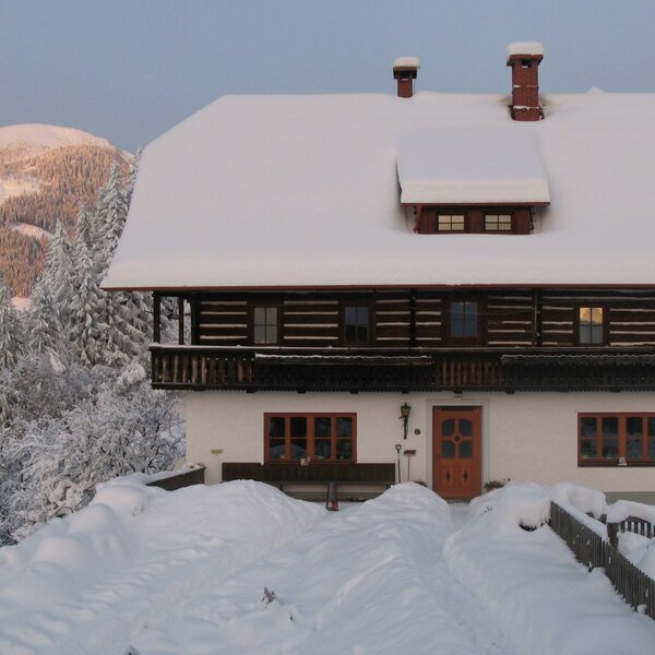 Der verschneite Bauernhof mit Holzbalkon und leuchtenden Fenstern, umgeben von schneebedeckten Bergen und Wäldern.