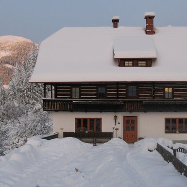 Der verschneite Bauernhof mit Holzbalkon und leuchtenden Fenstern, umgeben von schneebedeckten Bergen und Wäldern.